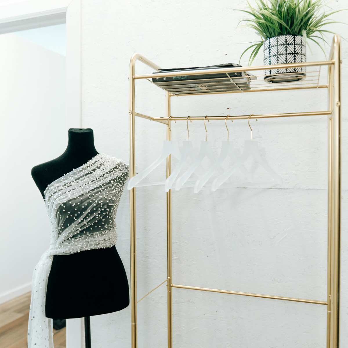 A black mannequin dressed with a sheer, embellished wrap stands next to a gold clothing rack with clear hangers and a potted plant on top.