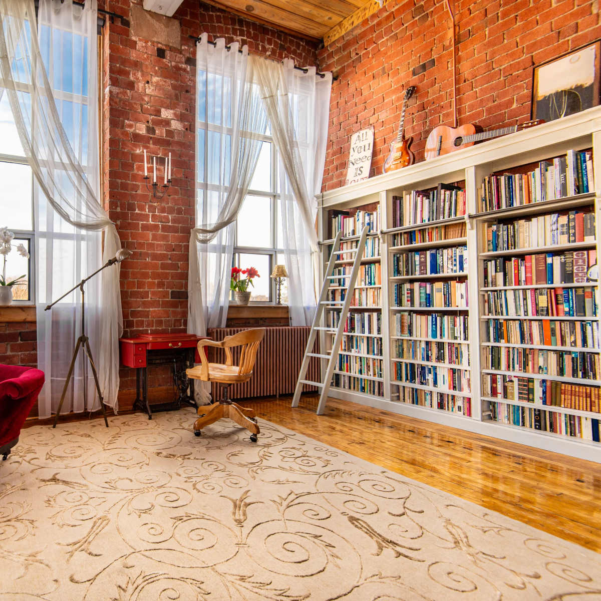 A cozy reading nook features a red velvet sofa, bookshelves filled with books, and large windows draped with sheer curtains in a brick-walled room.