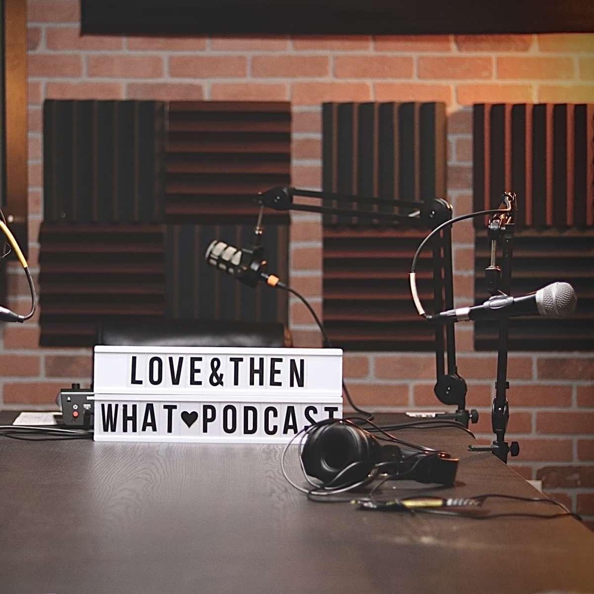 A podcast studio setup featuring two microphones, headphones, and a lightbox displaying the title "LOVE & THEN WHAT PODCAST" against a brick wall backdrop with acoustic panels.