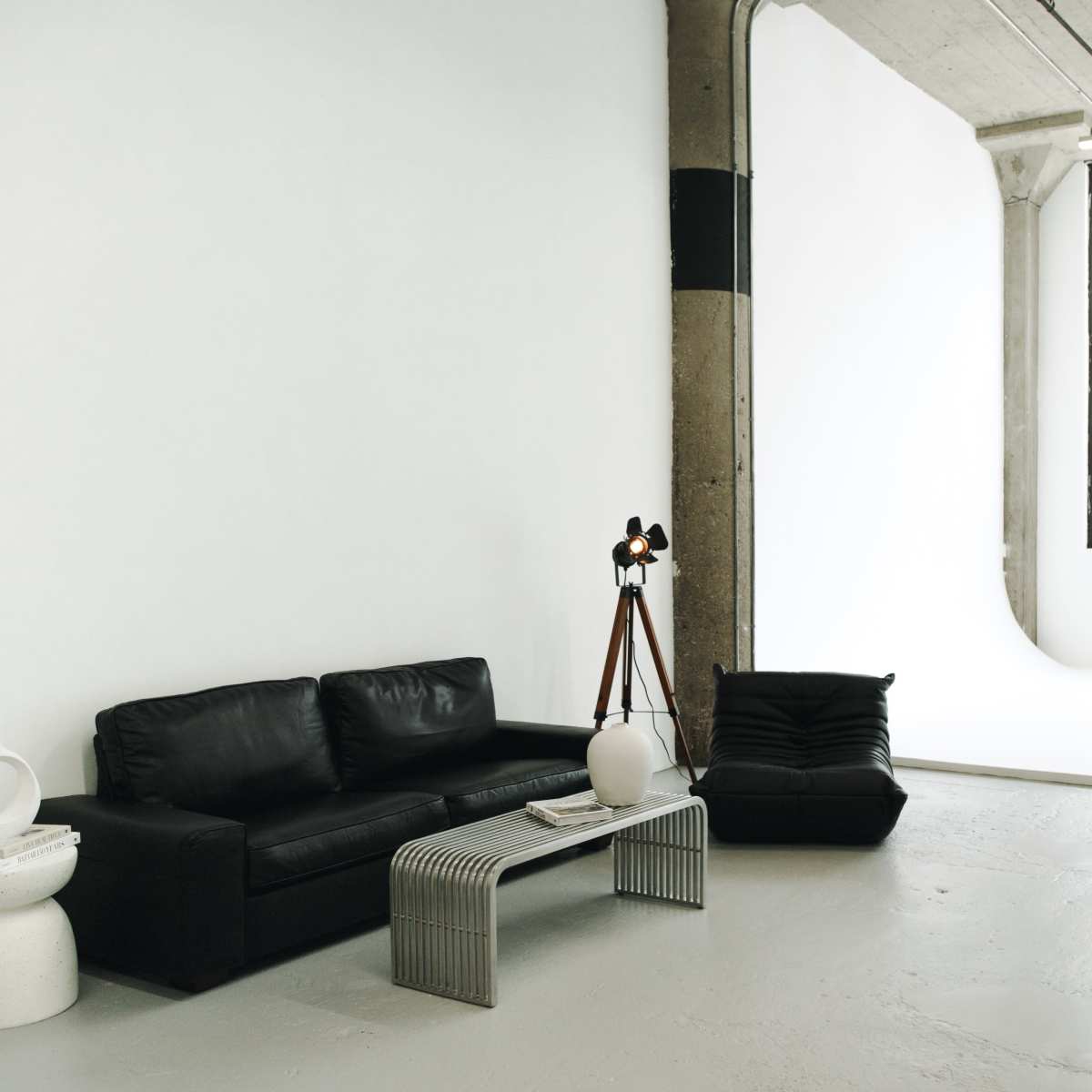 A spacious, well-lit interior featuring a black leather sofa, a modern table with a white vase, and large windows alongside a minimalist backdrop.