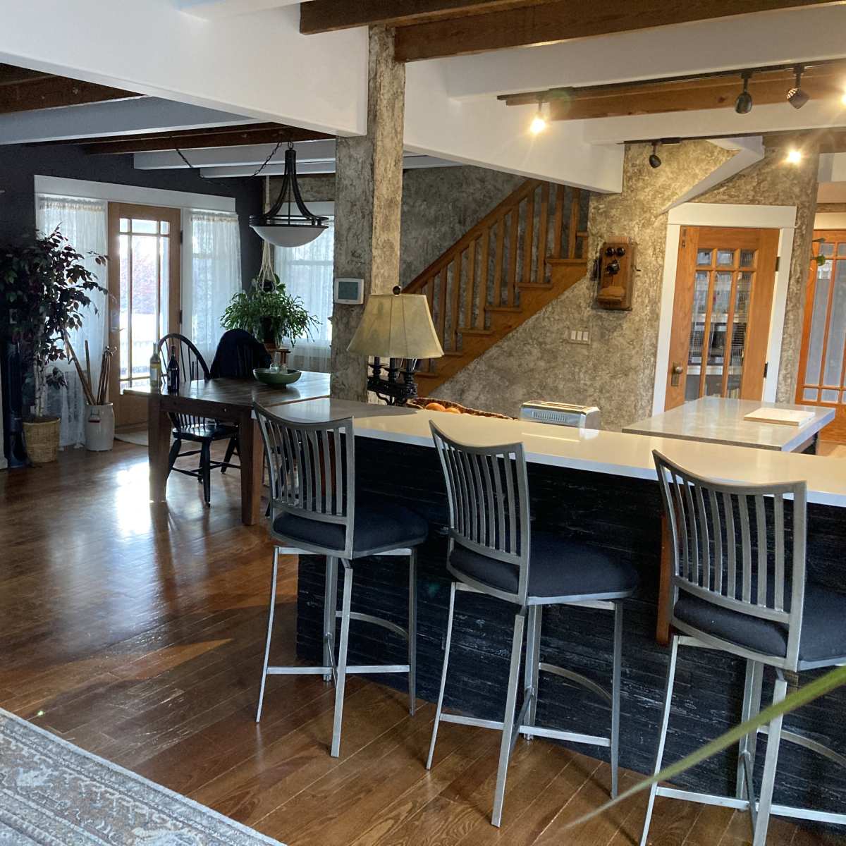A spacious living area with a dark sofa, an island bar with stools, a dining table, and a staircase leading to an upper level, illuminated by natural light through large windows.