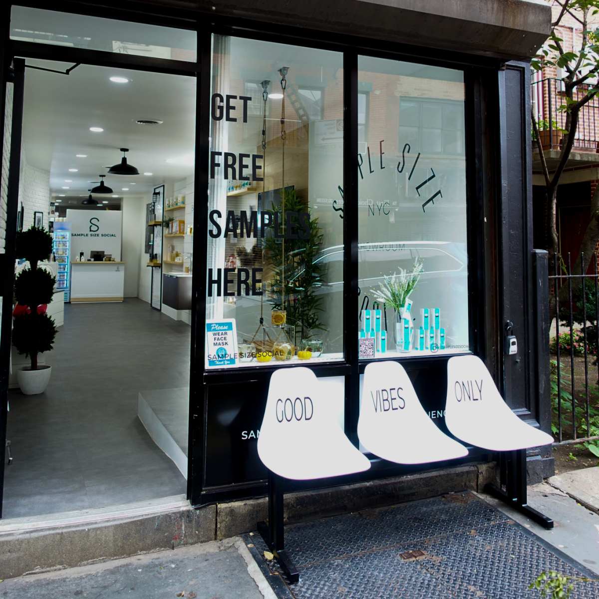 A storefront with large glass windows, featuring a sign that reads "GET FREE SAMPLES HERE" and three white benches labeled "GOOD VIBES ONLY" in front.