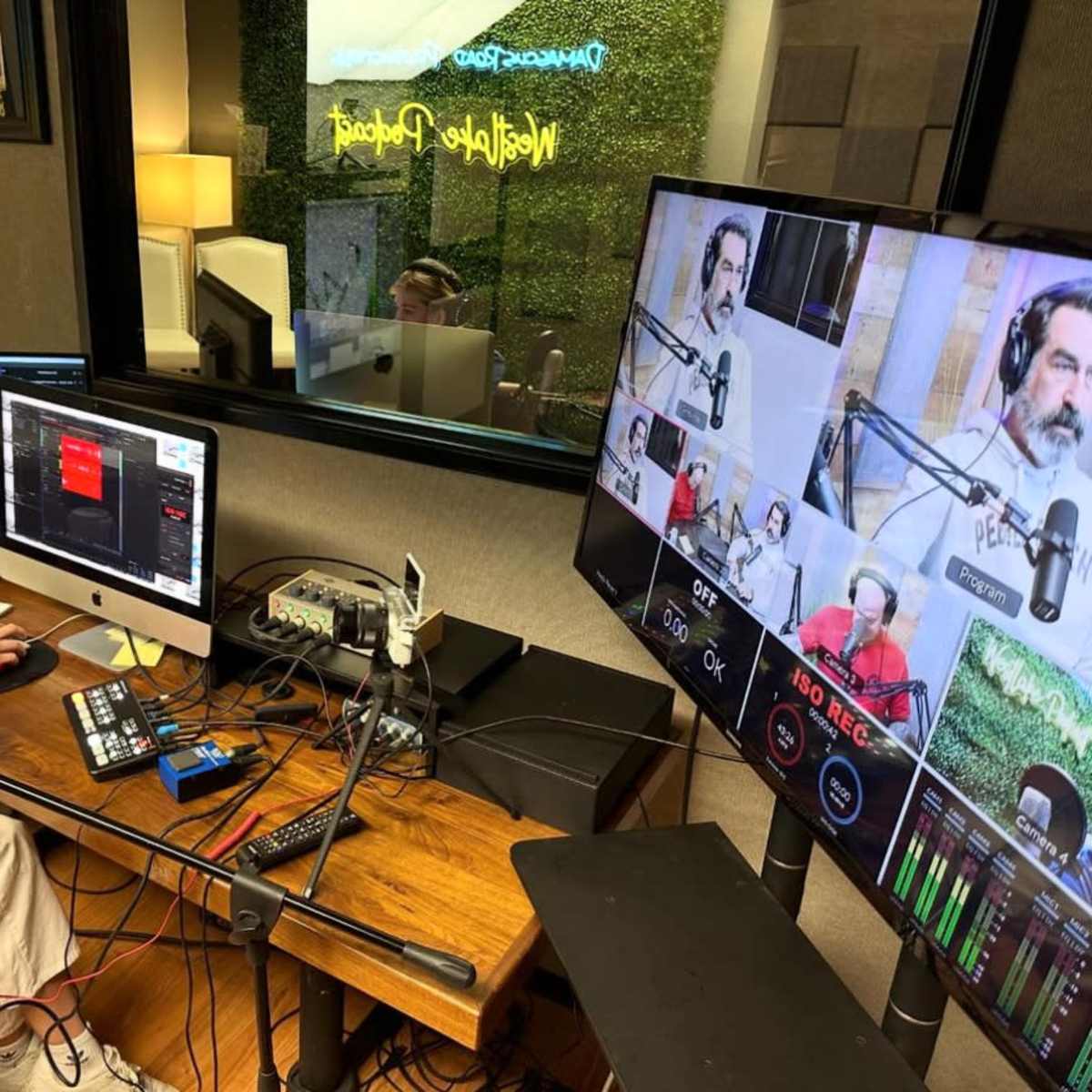 A person sits at a desk with multiple monitors and audio equipment, while engaging in a recording session in a well-lit studio.