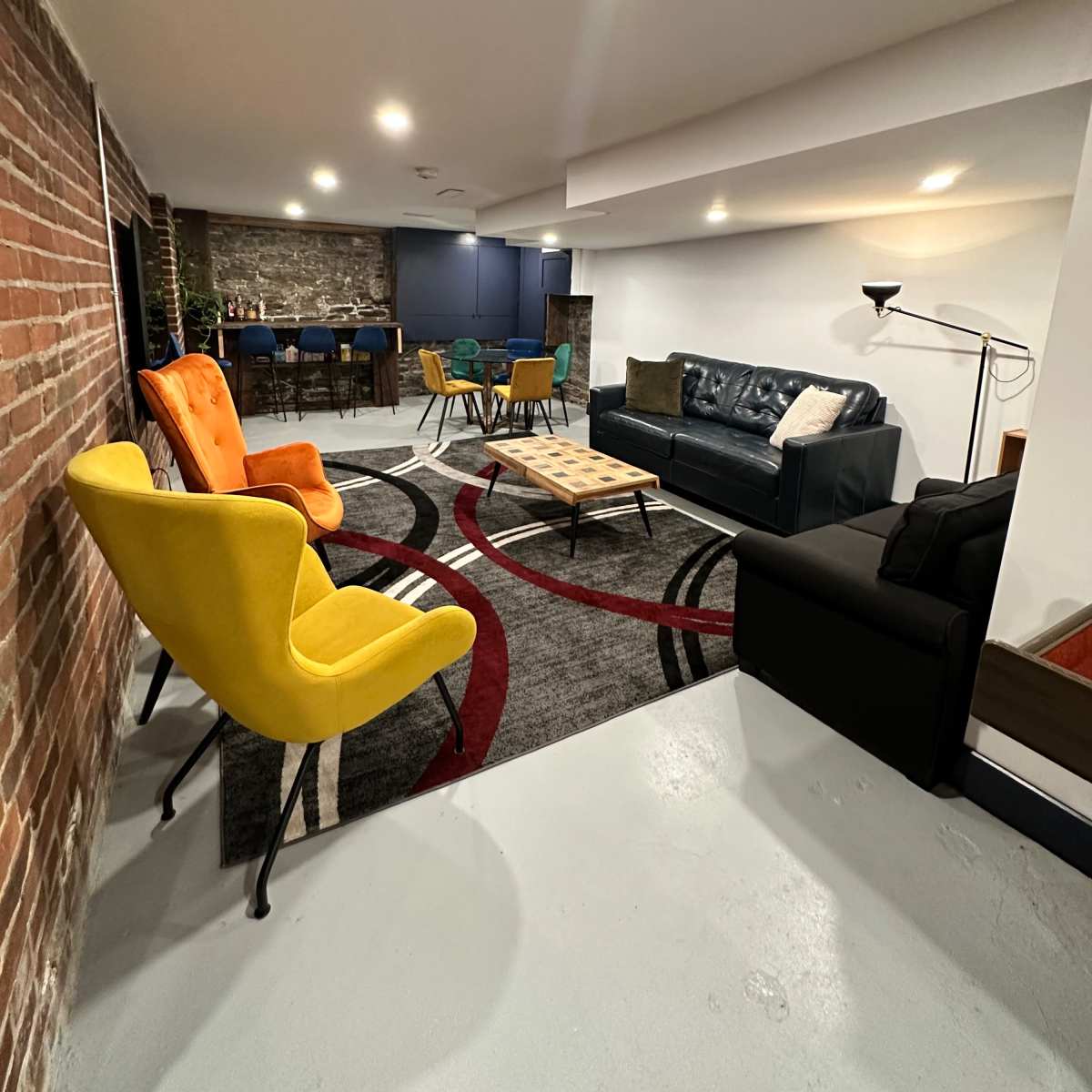 A cozy basement lounge area featuring colorful armchairs, a dark sofa, a coffee table, and exposed brick and stone walls.