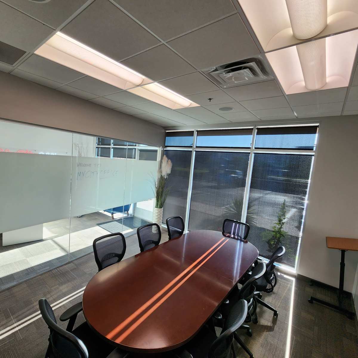 A well-lit conference room with a large, oval table surrounded by black chairs, featuring glass walls and windows that allow natural light to enter.