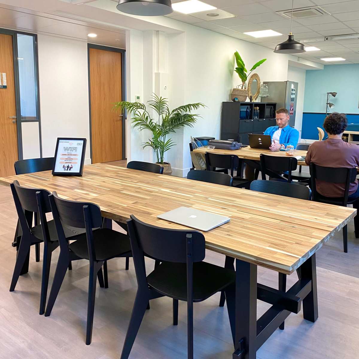 A modern office space with a large wooden table surrounded by black chairs, where two people are working on laptops.