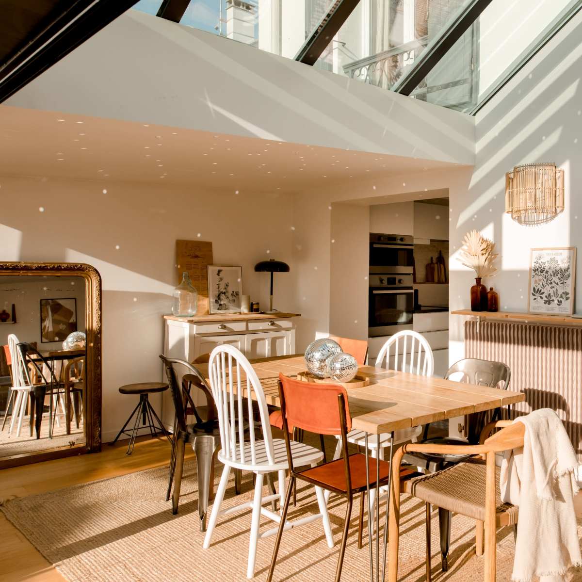 A bright, modern dining area features a table surrounded by various chairs, with large windows allowing daylight to create patterns on the wooden floor.