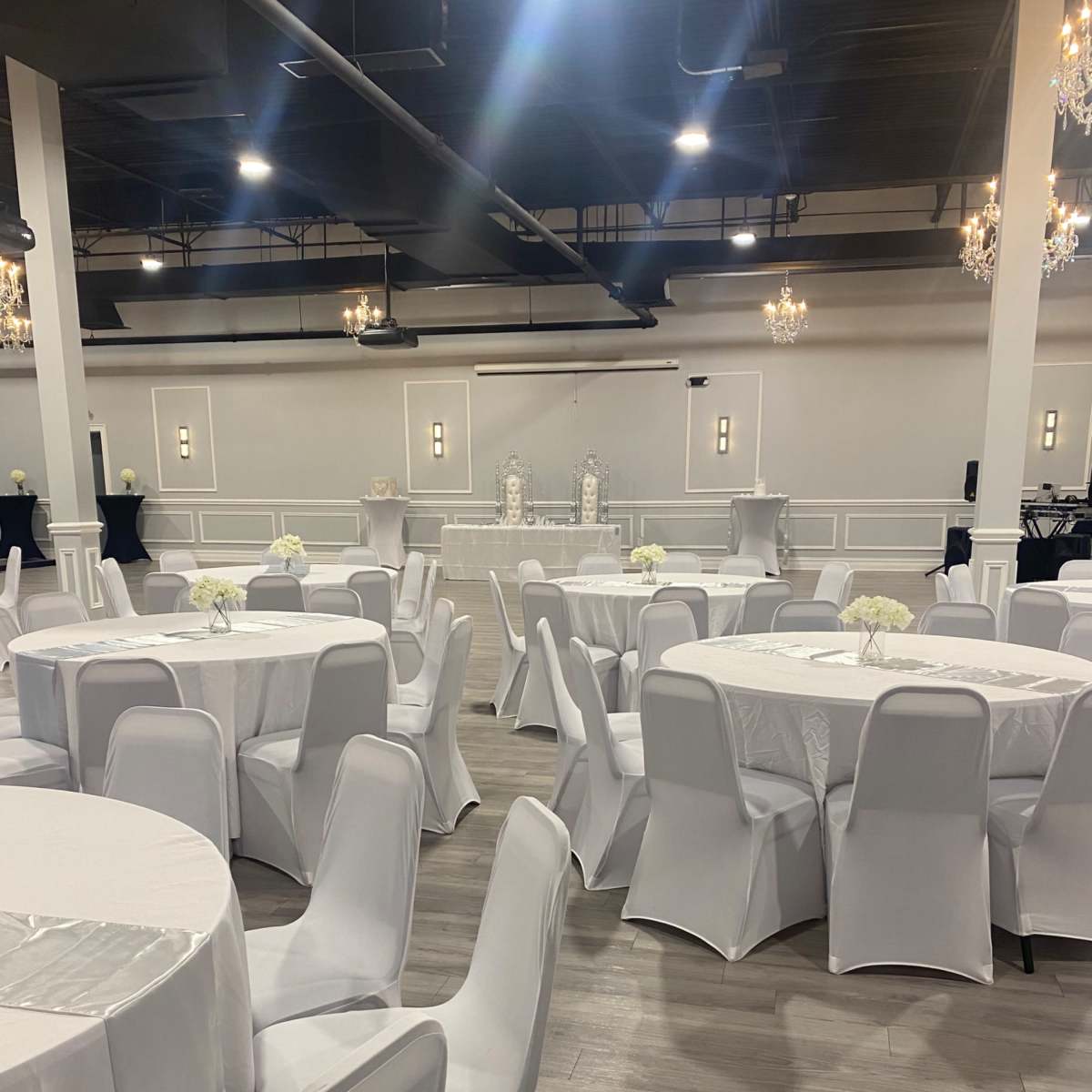 A large event space with round tables covered in white tablecloths and white chairs, arranged in a setup for an event, with chandeliers hanging above.