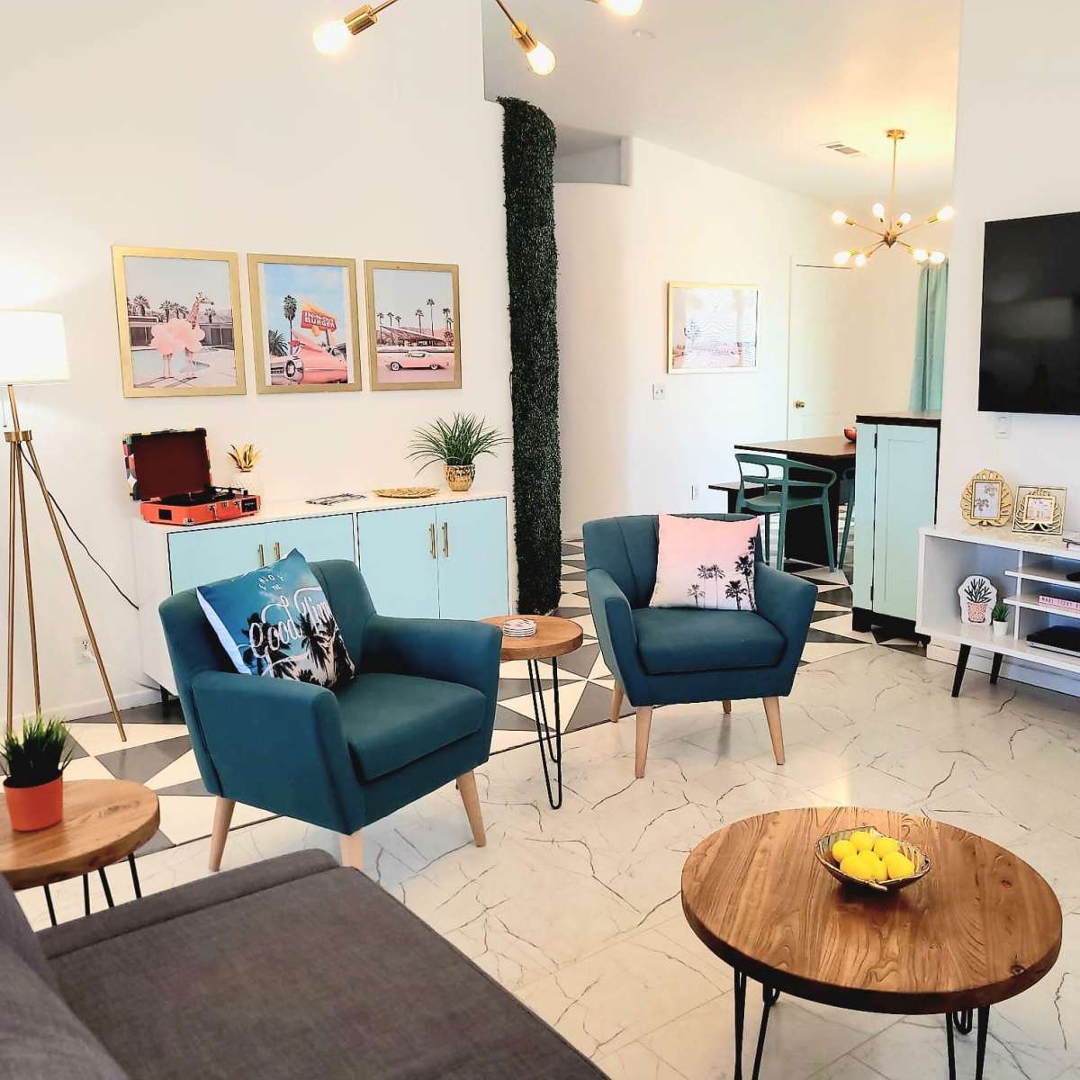 A modern living room with two teal chairs, a wooden coffee table, and a large television mounted on the wall, surrounded by decorative elements and art.
