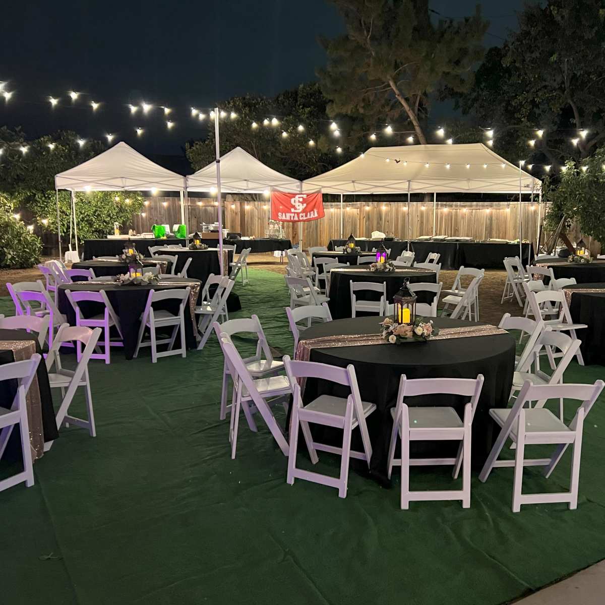 A nighttime outdoor event space set up with multiple tables and tents, adorned with string lights overhead.