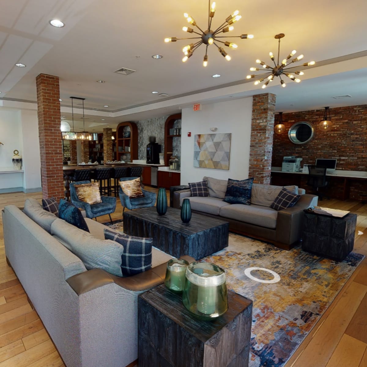 A modern lounge area with two gray sofas, wooden tables, a decorative rug, and exposed brick walls, creating a welcoming space.