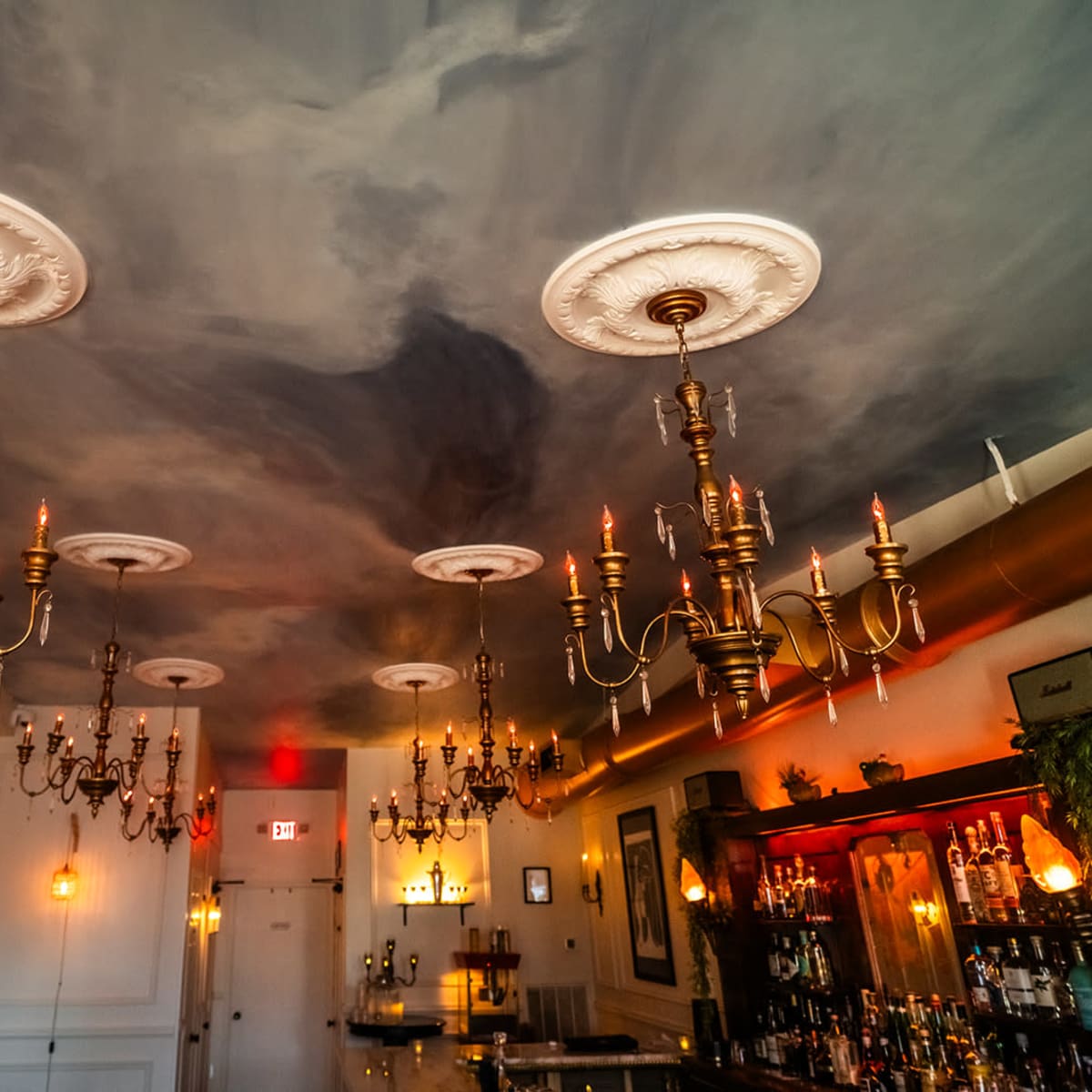 A dimly lit bar interior with ornate chandeliers hanging from a painted ceiling.