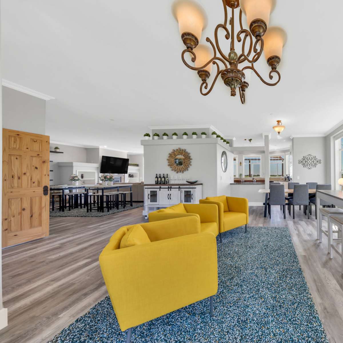 A brightly lit living area with two yellow armchairs, a patterned rug, a bar table with wine display, and an open view into a modern kitchen.