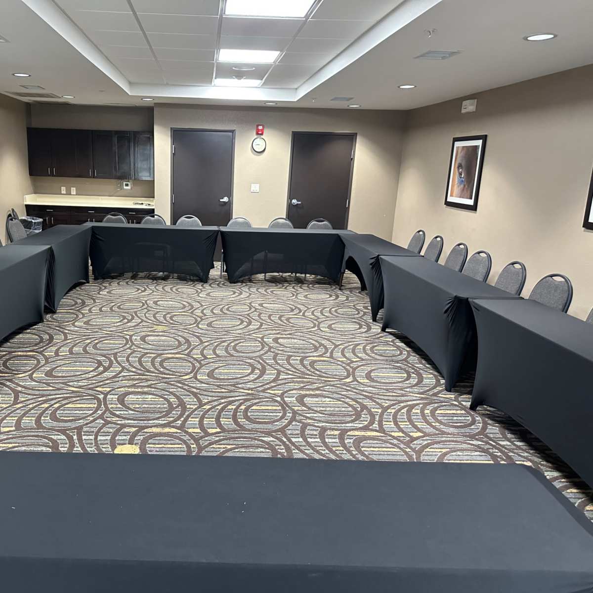 A rectangular meeting room arranged with black tables and chairs set in an oval configuration on a patterned carpet.