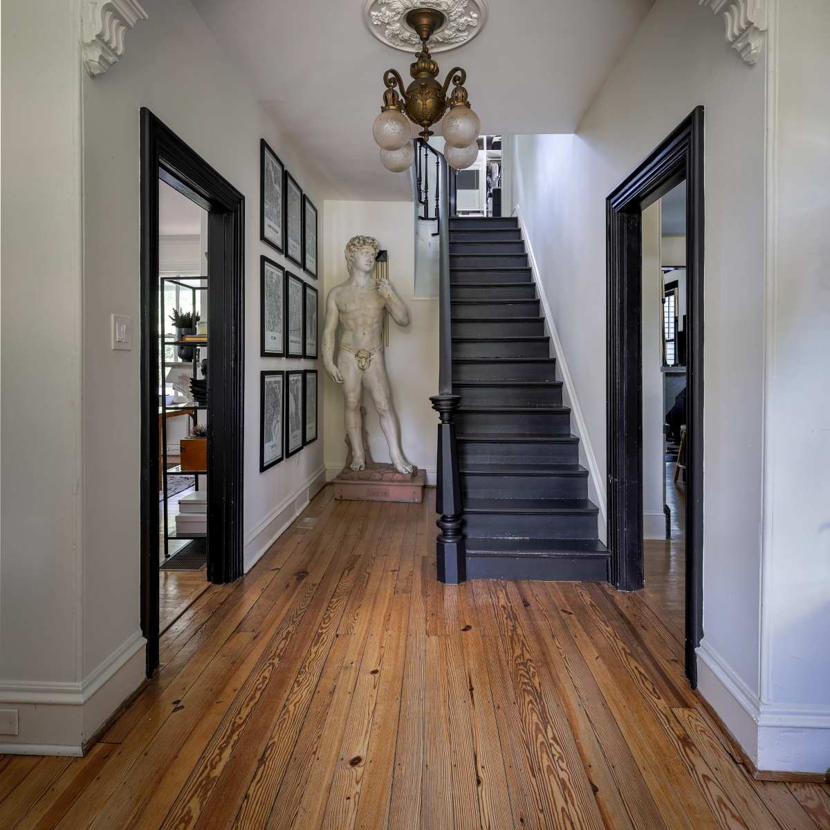 A hallway features a staircase ahead, flanked by black door frames and adorned with framed pictures on one side, while a statue stands near the wall.