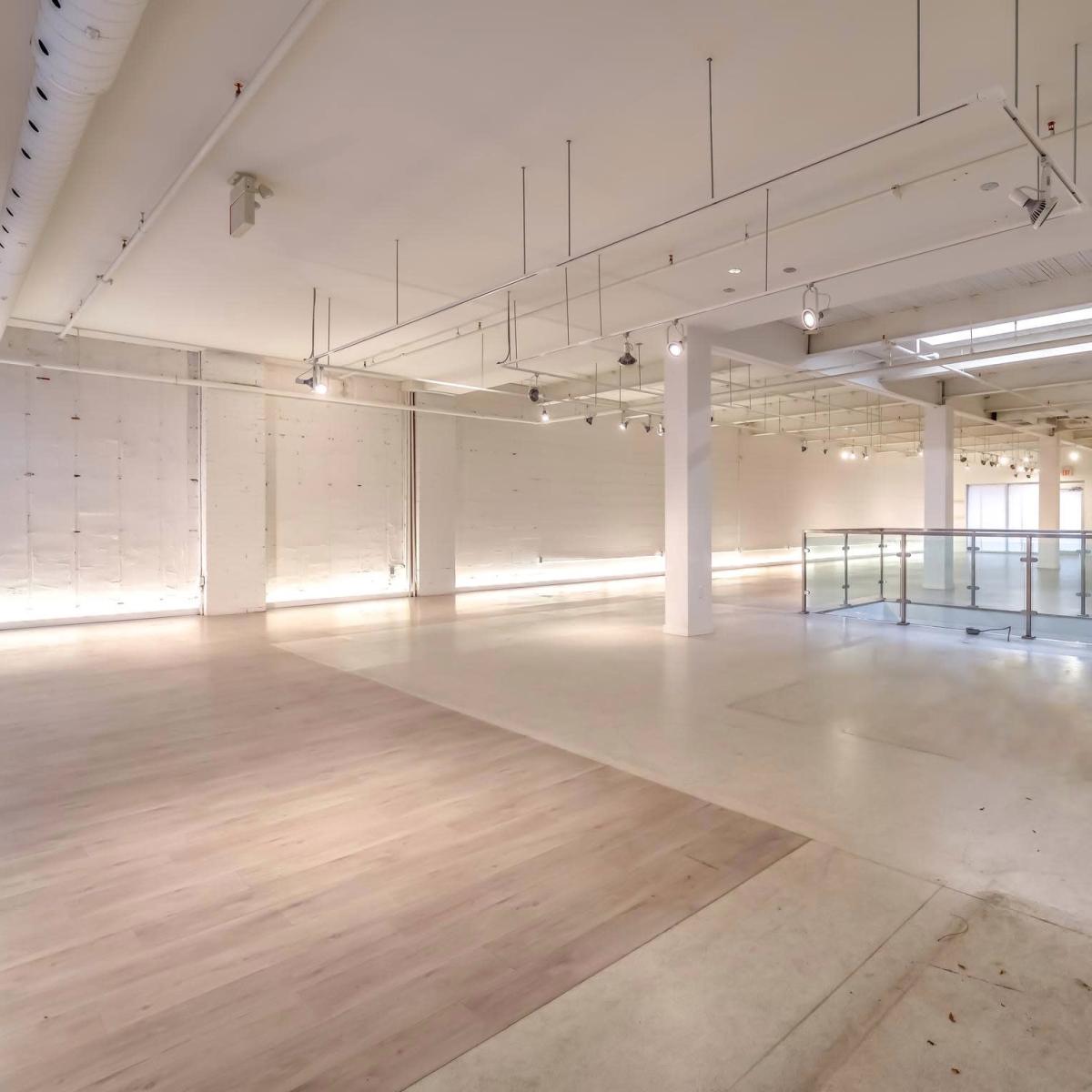 An empty, well-lit interior space with polished floors and large windows, featuring a mezzanine level with a glass railing.