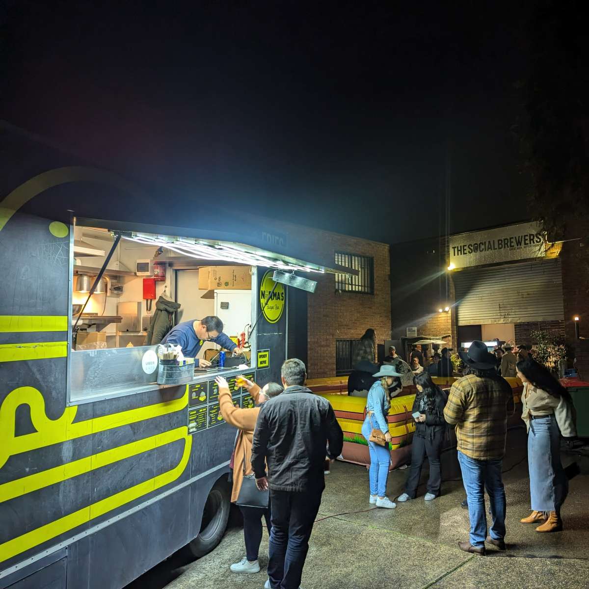 A food truck is serving a line of customers outside a brewery at night.