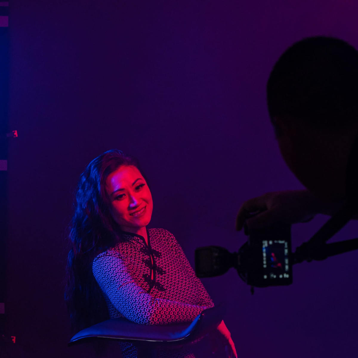 A woman sits in a chair as a photographer takes her picture in a dimly lit studio with colorful lighting.