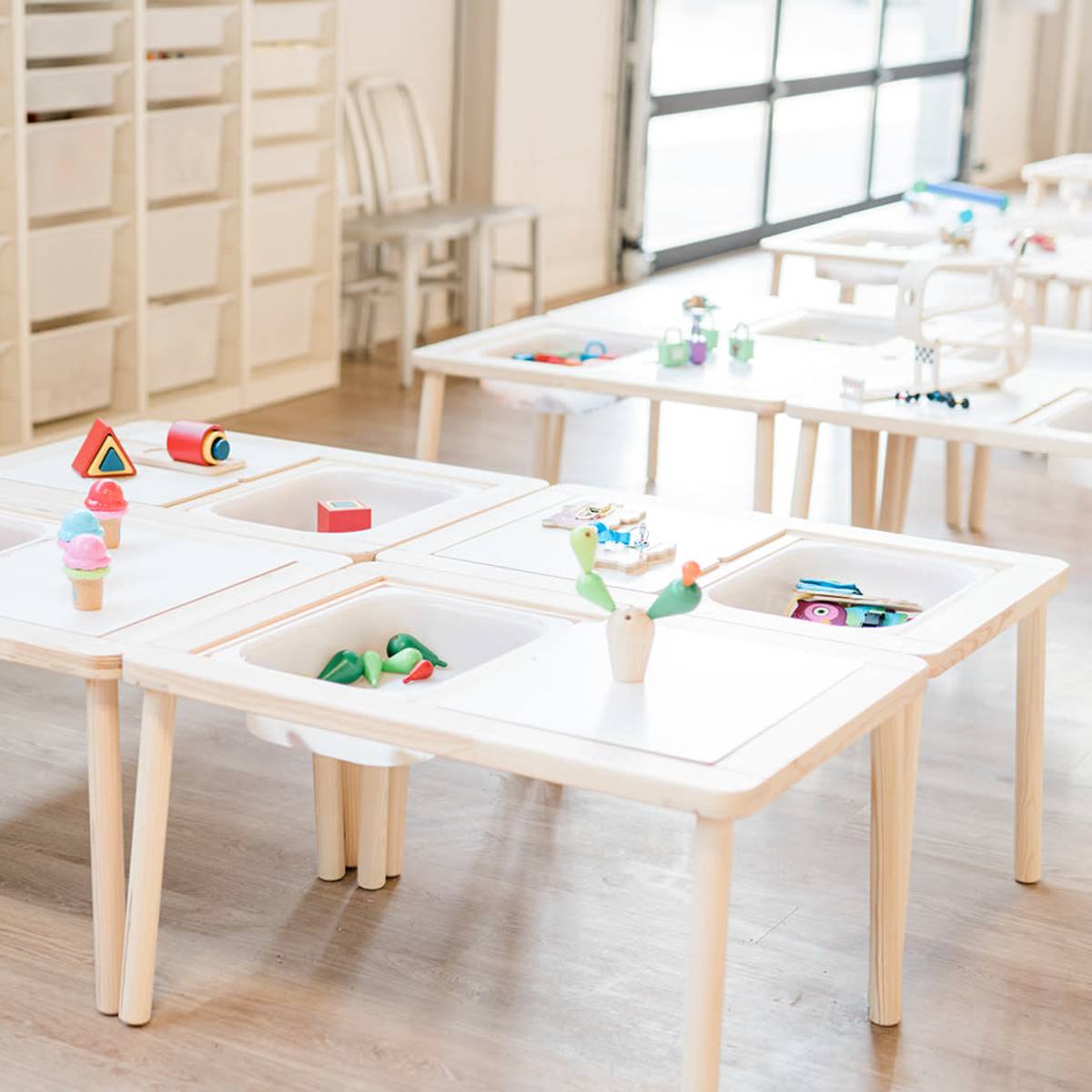 A classroom filled with white tables, each with various colorful educational toys and materials arranged in their center.