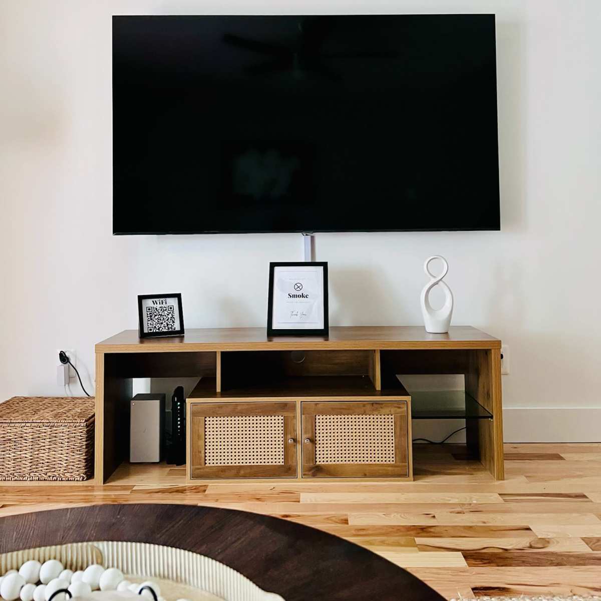 A minimalist living room setup with a large flat-screen TV mounted on the wall above a wooden TV stand, which features decorative items and storage baskets.