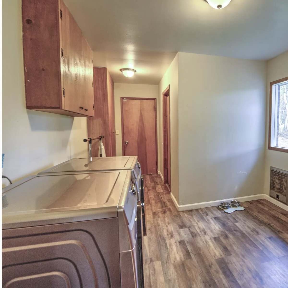A laundry room with a washer and dryer on the left, a wooden door leading outside, and a window on the right allowing natural light to enter.