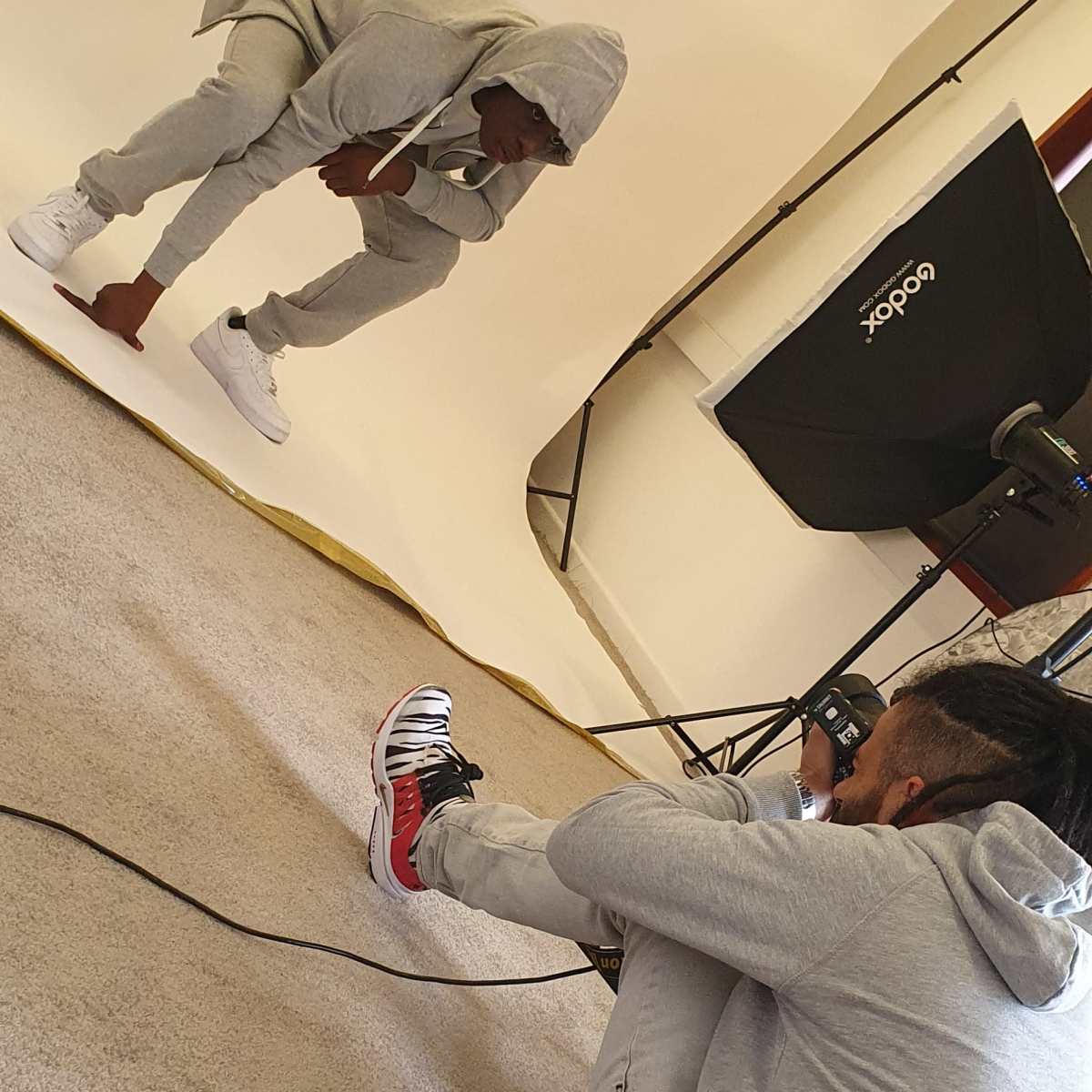 A photographer captures a seated model in a gray tracksuit against a neutral backdrop in a studio setting.