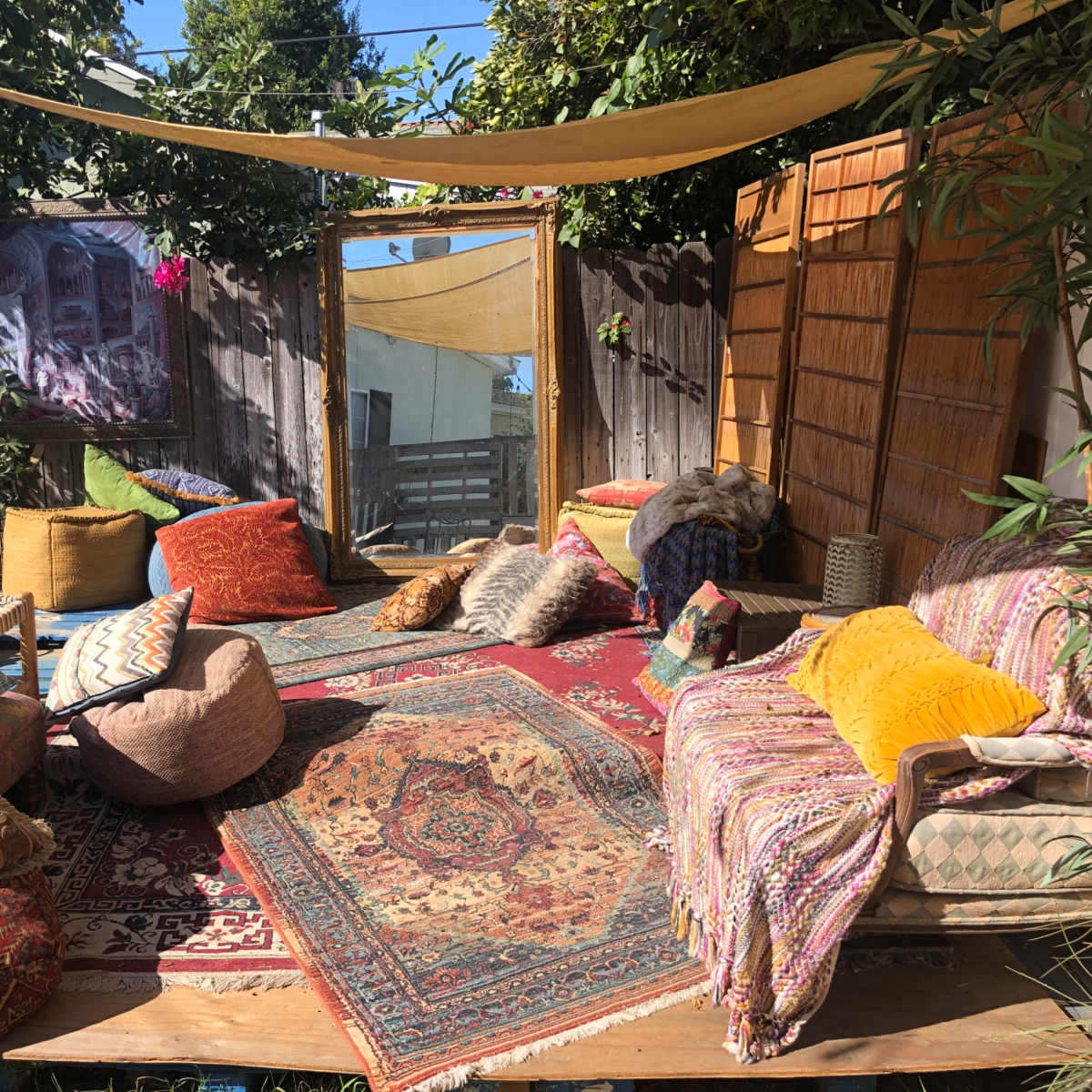 The scene features a cozy outdoor seating area adorned with various cushions, rugs, and a large mirror surrounded by greenery.