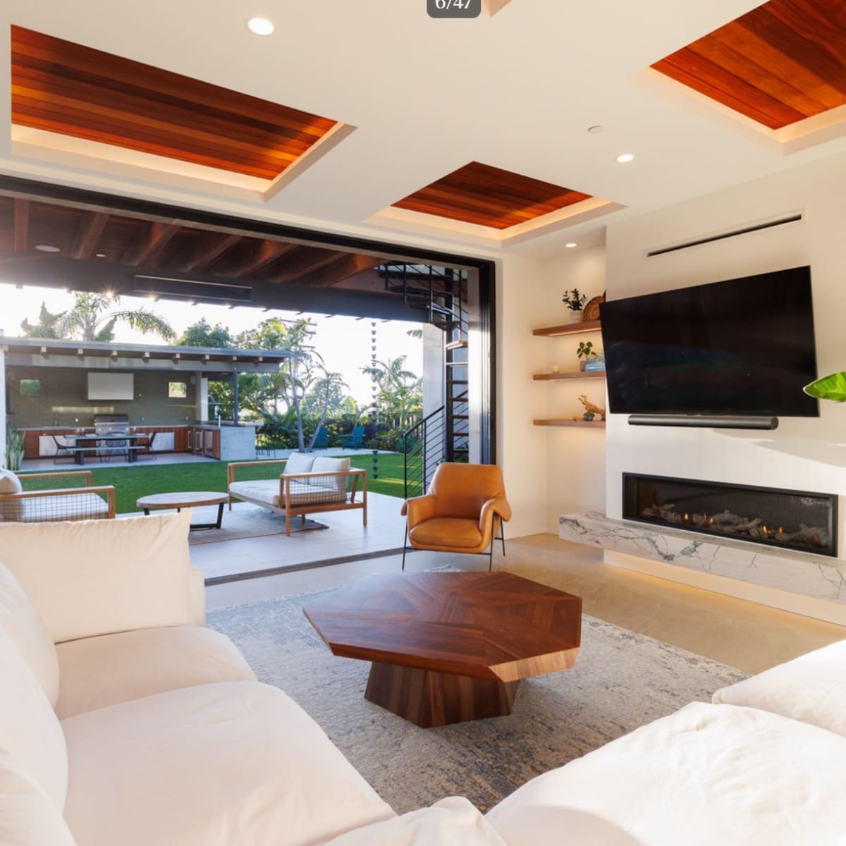 The modern living room features a large sectional sofa, a wooden coffee table, and a flat-screen TV mounted on the wall, with wide doors opening to a backyard patio.