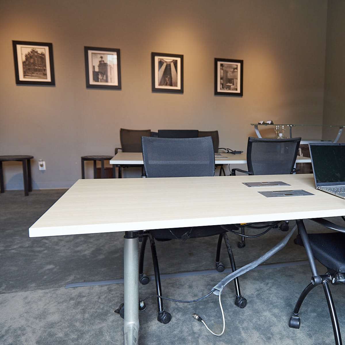 An office space with several desks and chairs arranged around a light-colored table, featuring a laptop and framed photographs on the walls.