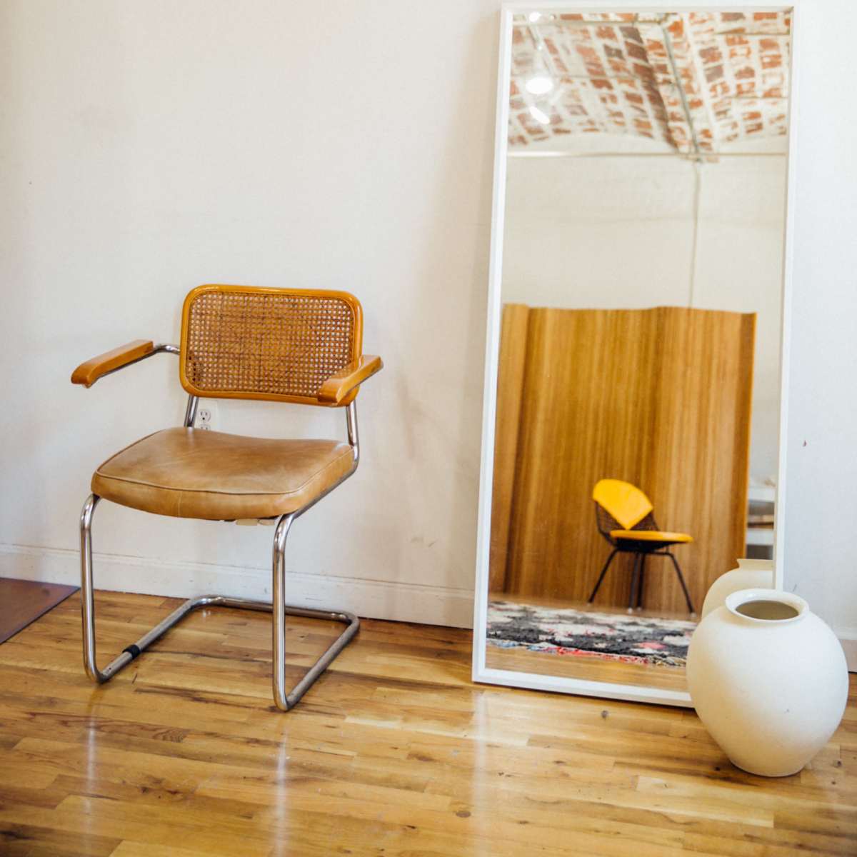 A chair with a woven backrest stands beside a large mirror reflecting a wooden panel and another chair in a minimalist interior space.