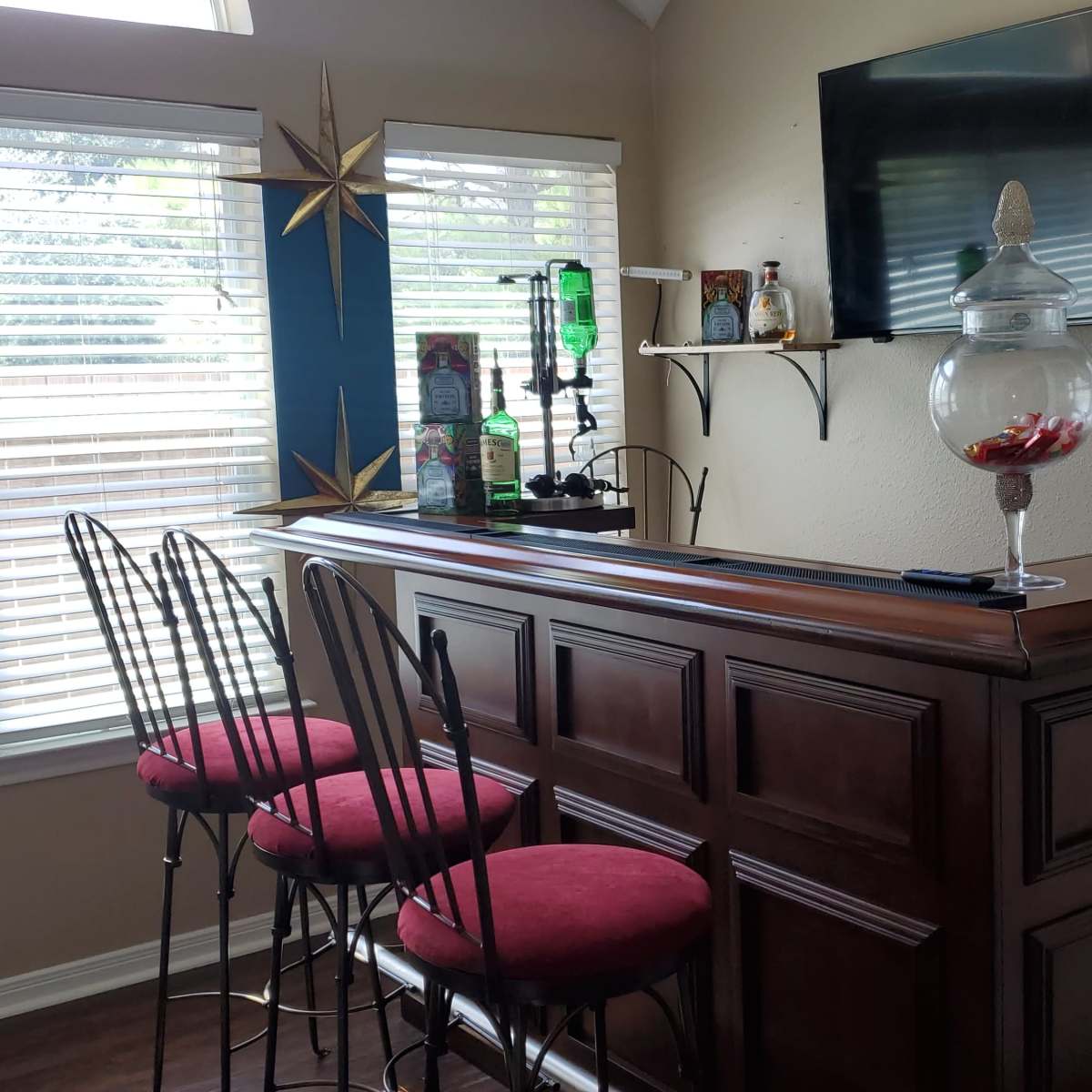 A home bar with four tall chairs, a wooden bar counter, and a wall-mounted television, surrounded by windows with white blinds.