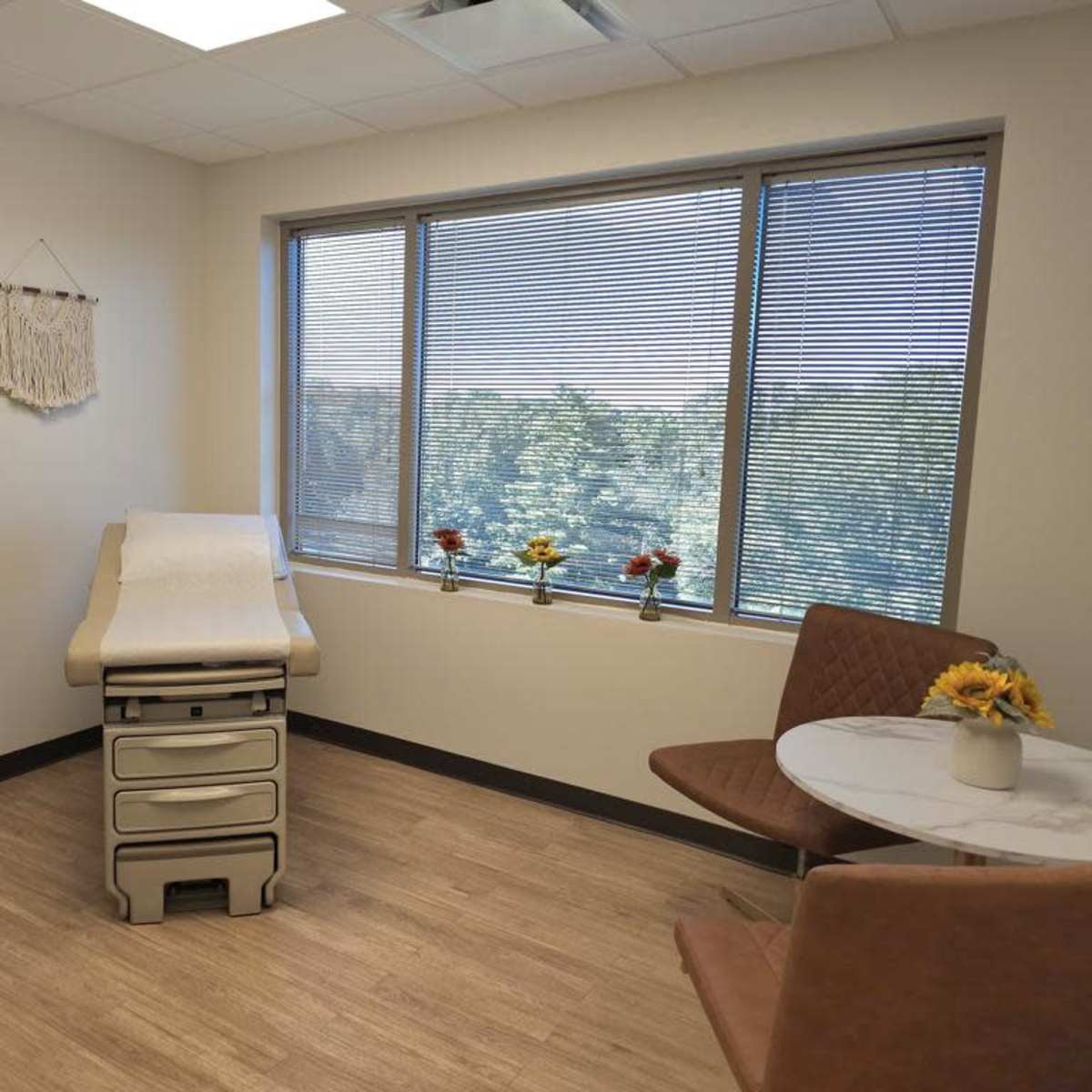 A medical examination room featuring an examination table, a small round table with two chairs, and a window with a view of greenery outside.