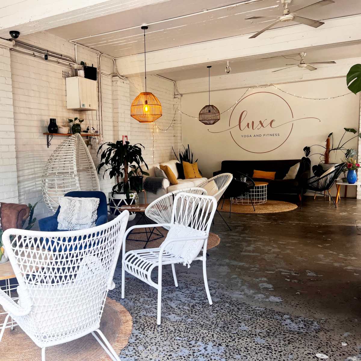A cozy fitness and yoga studio with comfortable seating, decorative woven light fixtures, and plants in a bright, airy space.