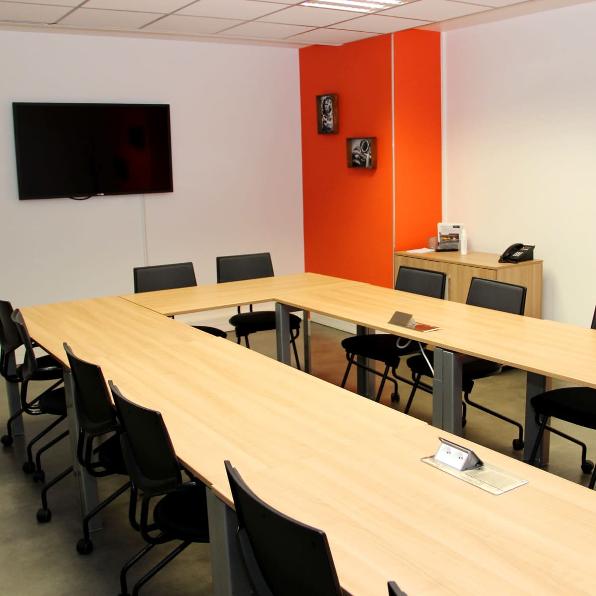 A rectangular conference room features a large wooden table surrounded by black chairs, equipped with a wall-mounted TV and a phone.