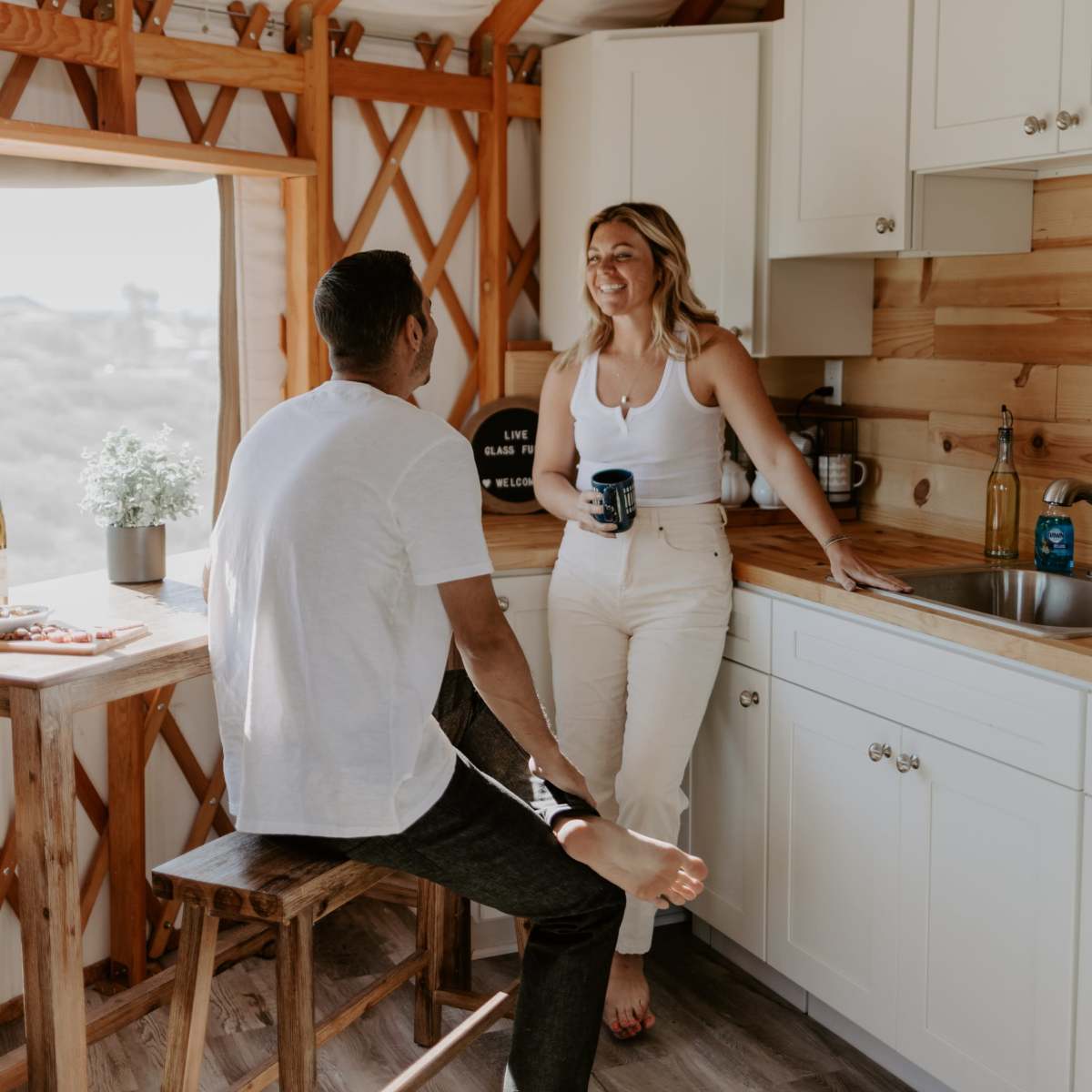 A man and a woman engage in conversation while standing in a rustic kitchen with wooden walls and white cabinetry.