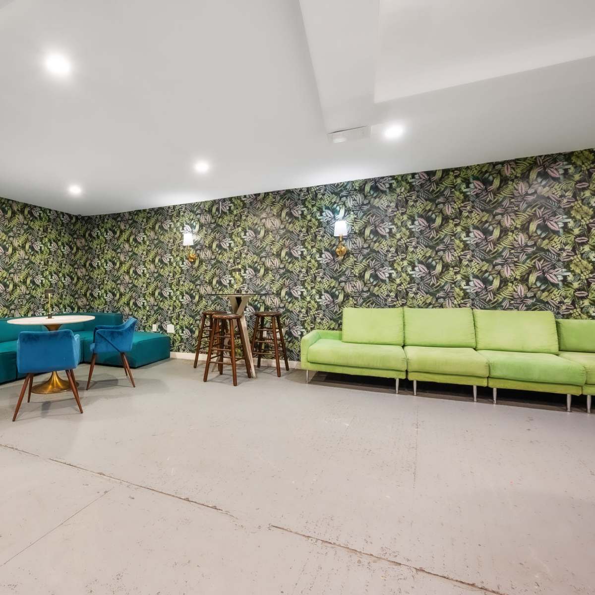 A modern room with a patterned green and tropical wallpaper, featuring a turquoise sectional sofa, a blue chair and table, and a green sofa along with two wooden bar stools.