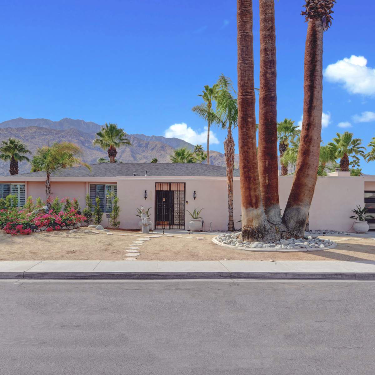 A single-story residential home with palm trees and a desert landscape sits at the end of a sidewalk, against a backdrop of mountains.
