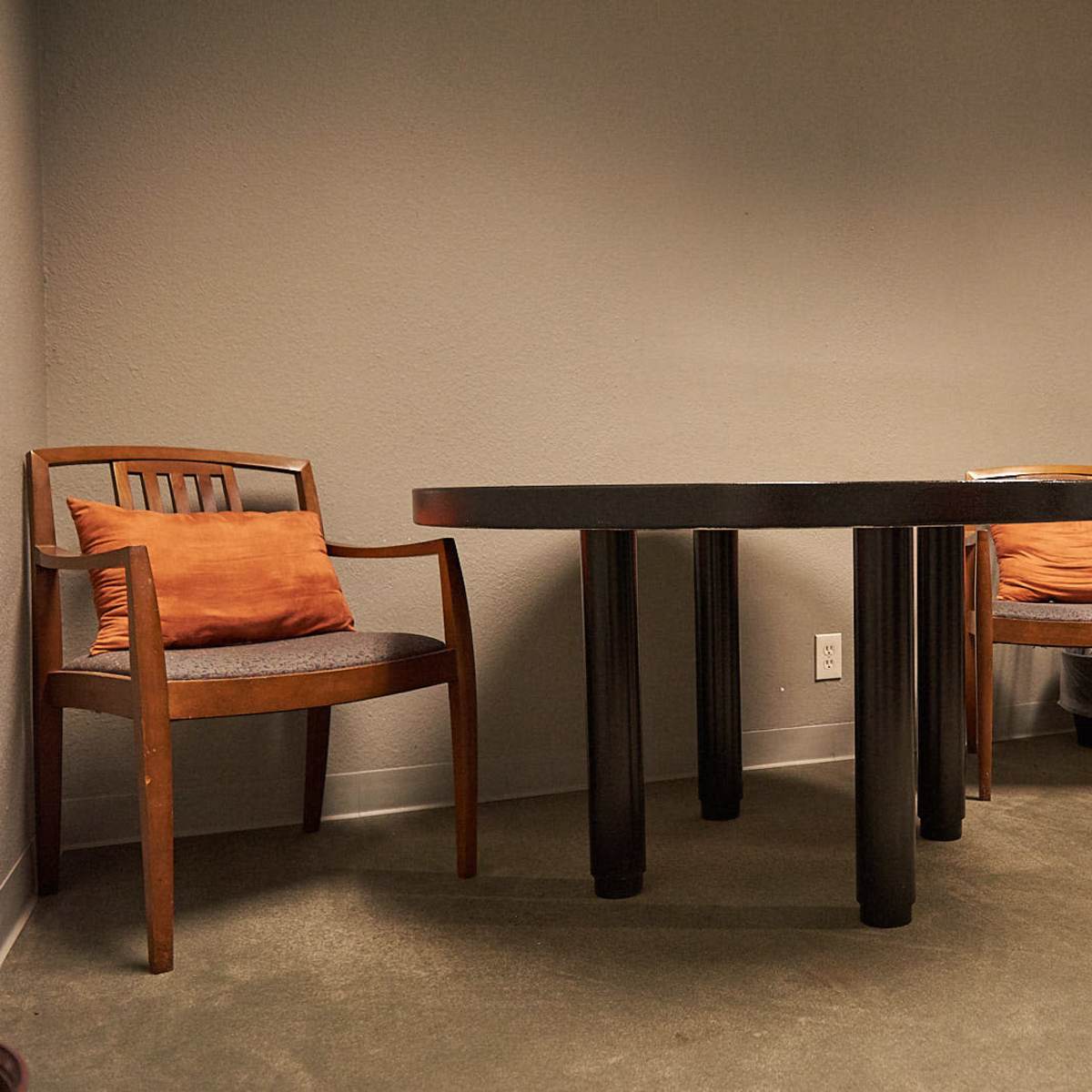A small room features a black table with four legs, two wooden chairs with orange cushions, and a television on a dresser against the wall.