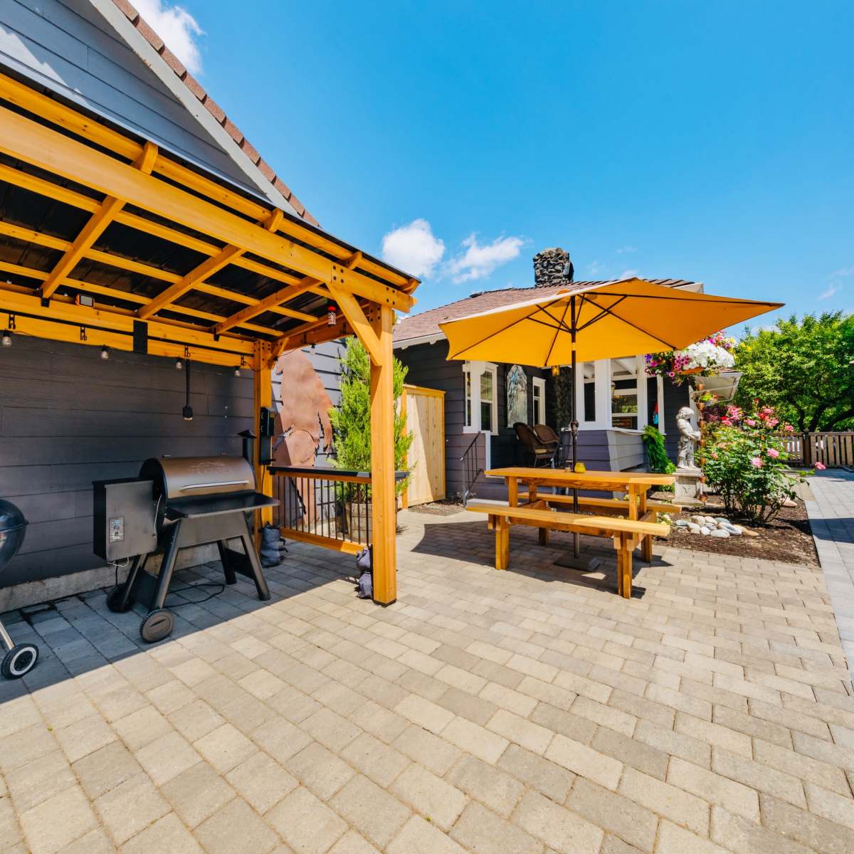 A patio area with a grill, a picnic-style table, and yellow umbrellas, surrounded by a well-maintained yard and a house in the background.