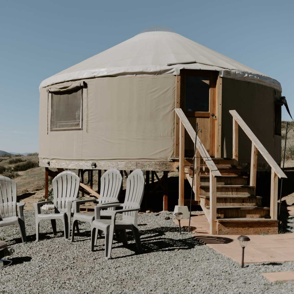 A yurt with a wooden deck and steps is surrounded by gravel and several plastic chairs.
