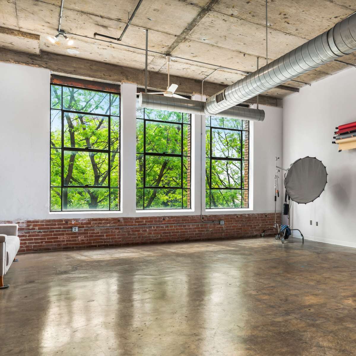 A spacious studio features large windows with views of green trees, exposed concrete ceilings, and photography equipment along the walls.