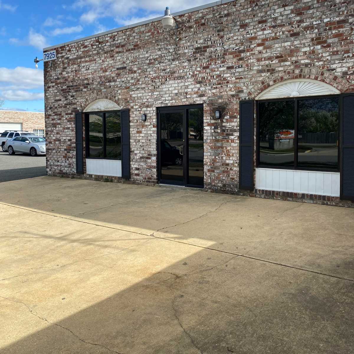 A brick building with large glass doors and window shutters, located on a paved area under a blue sky with scattered clouds.