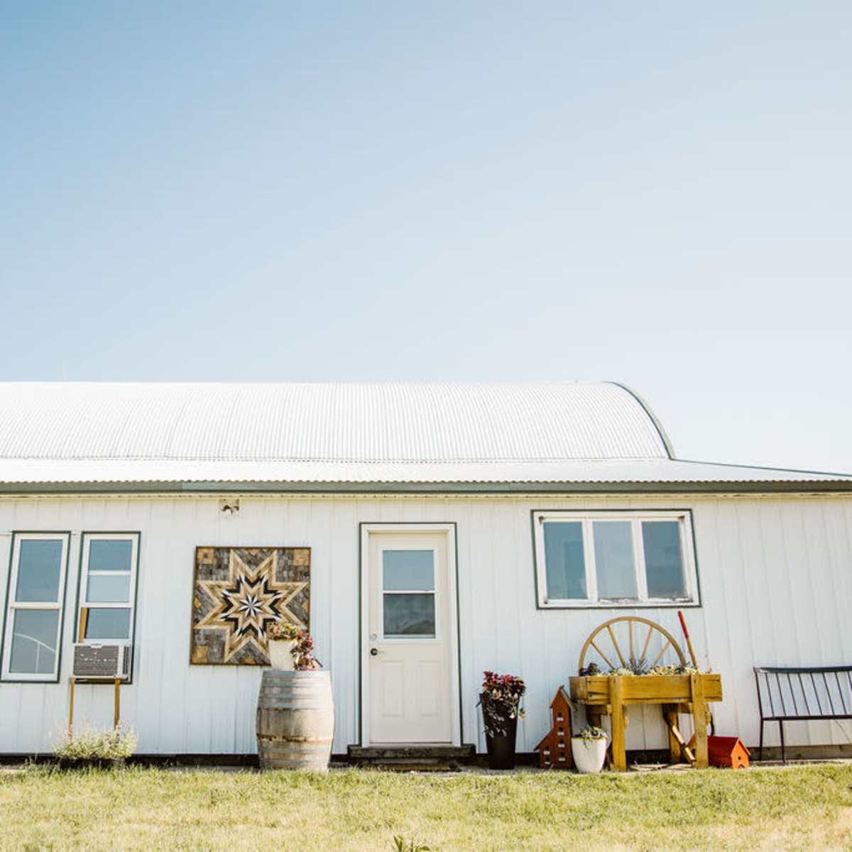 A white metal-clad farmhouse features large windows, a decorative star on the wall, and a yellow cart by the entrance.