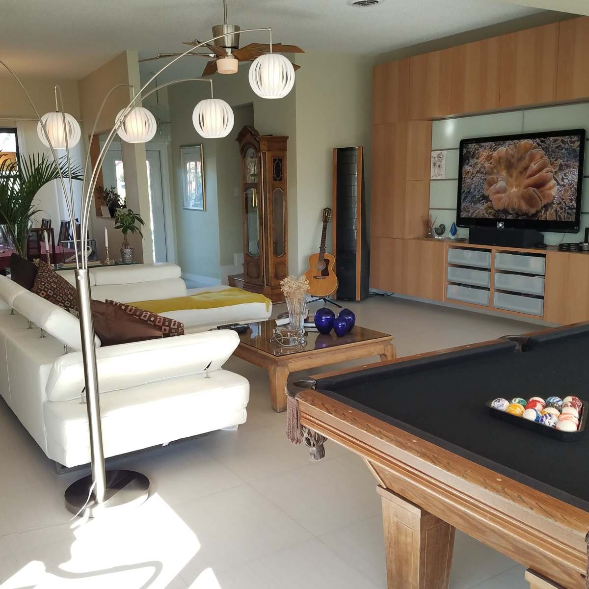 A spacious living area featuring a white sectional sofa, a wooden pool table, a large television mounted on a wall unit, and decorative plants.