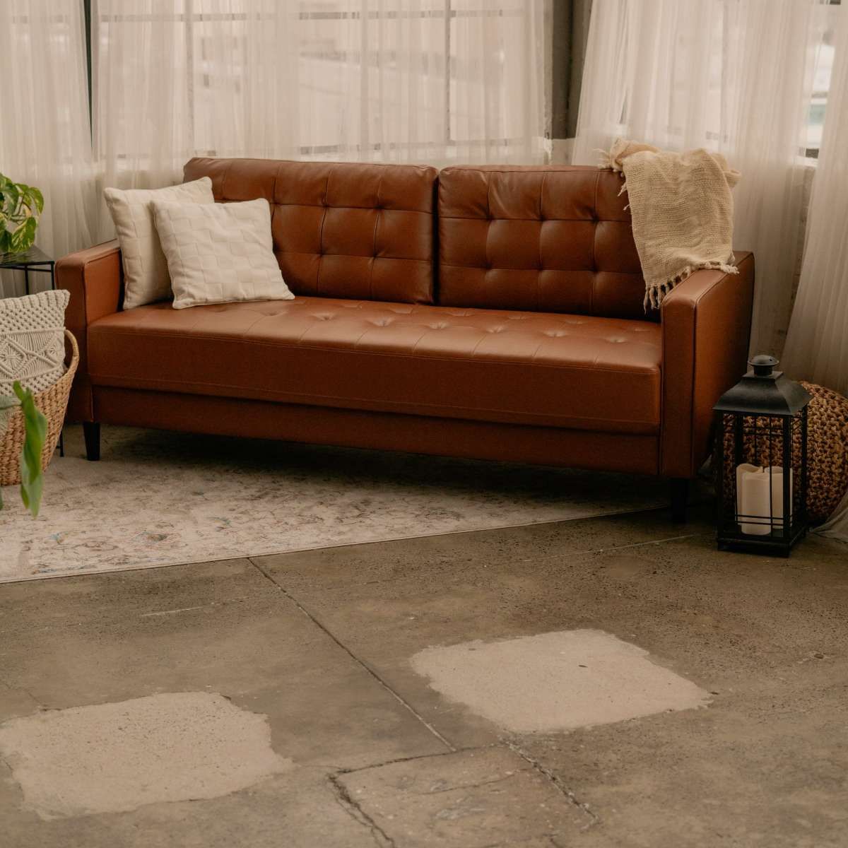 A brown leather couch is positioned in a bright, airy room with sheer curtains and a concrete floor.