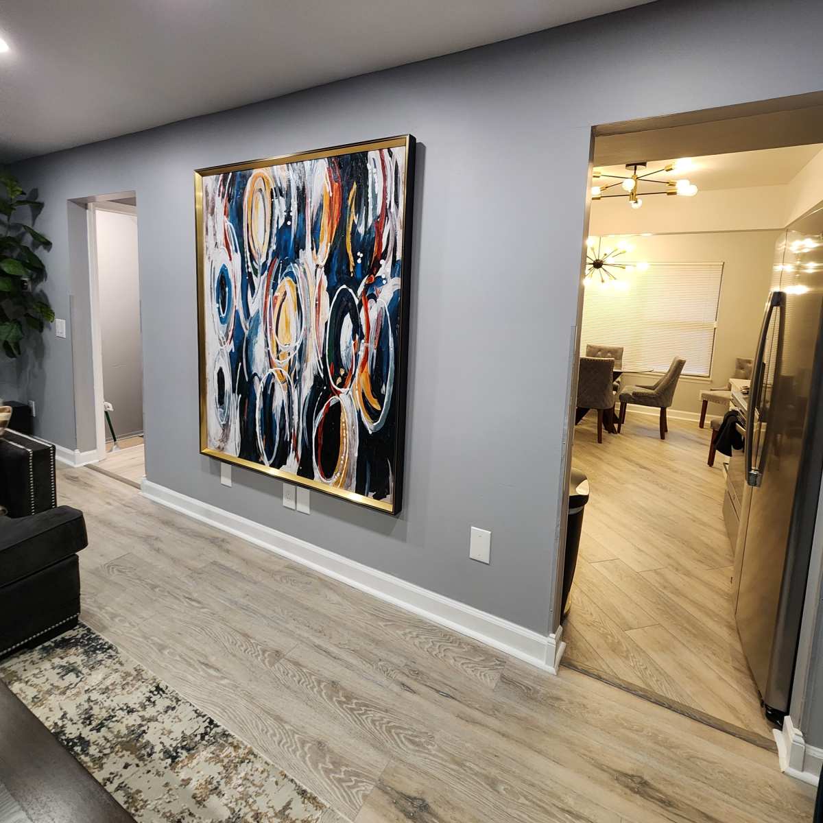 A large abstract painting with circular patterns is displayed on a gray wall, leading towards an open doorway that reveals a dining area.