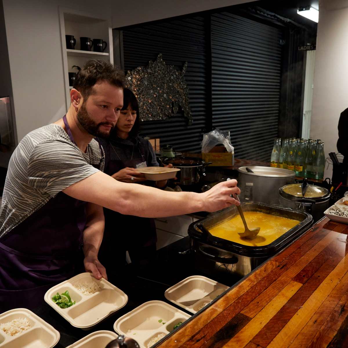 A man and a woman serve food from a large pot into disposable containers at a kitchen counter.