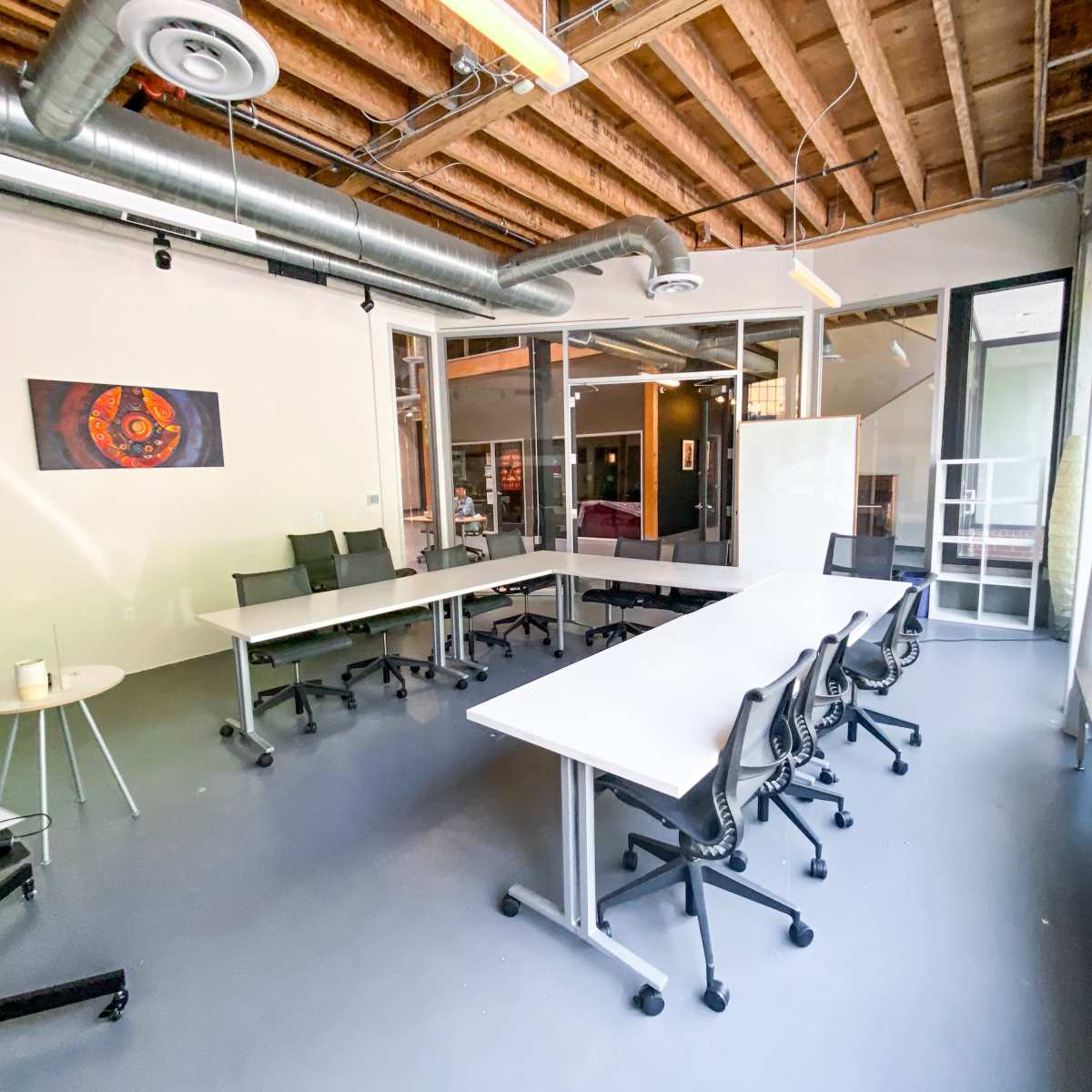 A modern conference room features a large table surrounded by chairs, with windows allowing natural light and a piece of artwork on the wall.