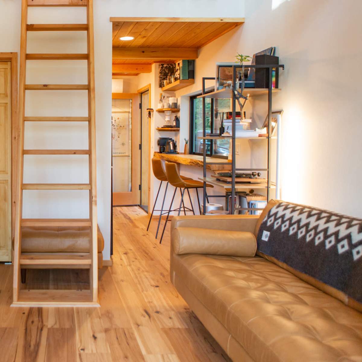A small living space featuring a wooden ladder leading to an upper area, a desk with a computer, a sofa, and a kitchen area with stools.