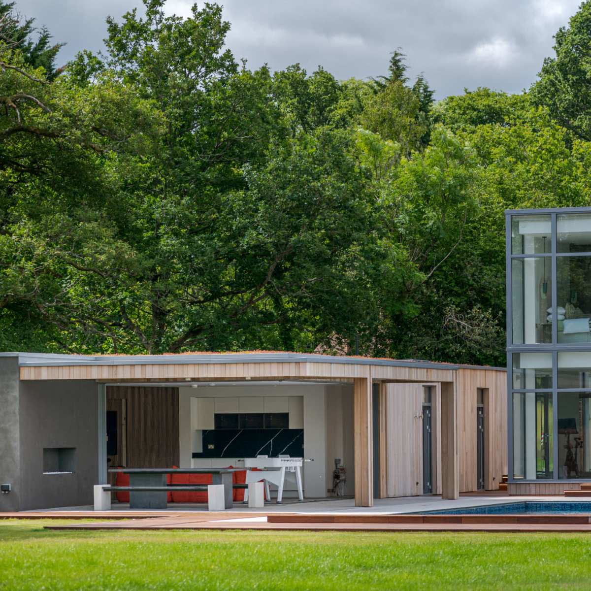 A modern house with large glass windows, a wooden exterior, and a swimming pool surrounded by a grassy area and lush trees.